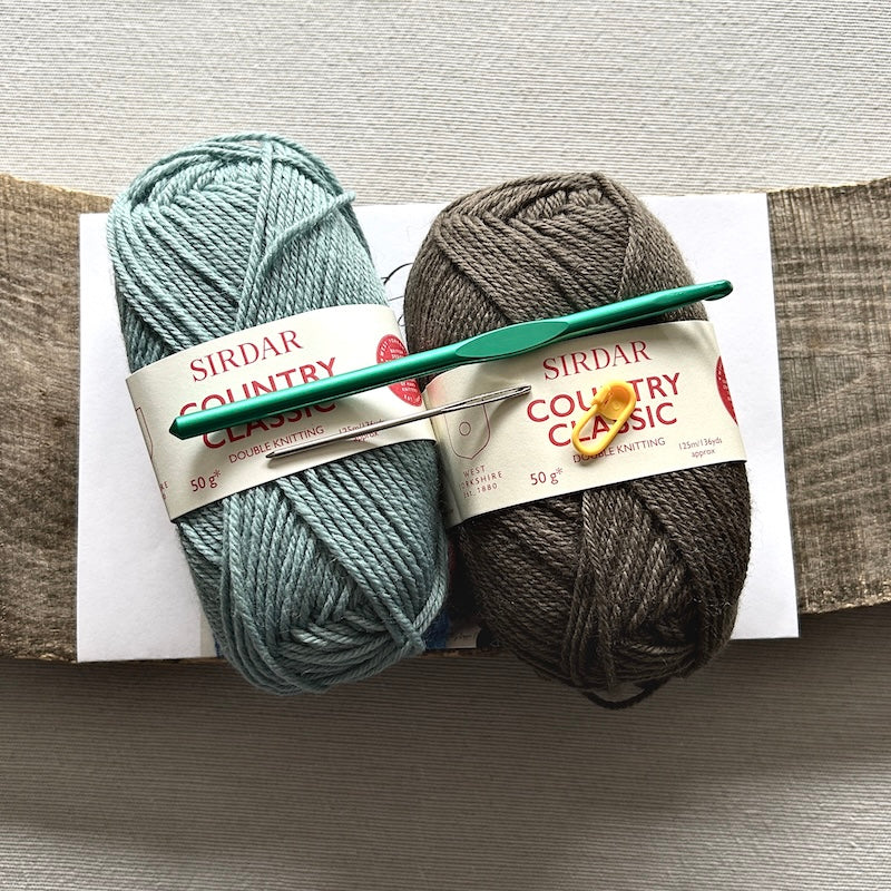 2 balls of yarn, a crochet hook and a pattern are sitting on a wooden board.