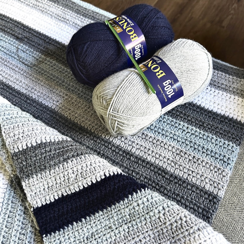 The finished couch throw, made from this crochet kit, is draped over a couch with 2 balls of yarn and a crochet hook.