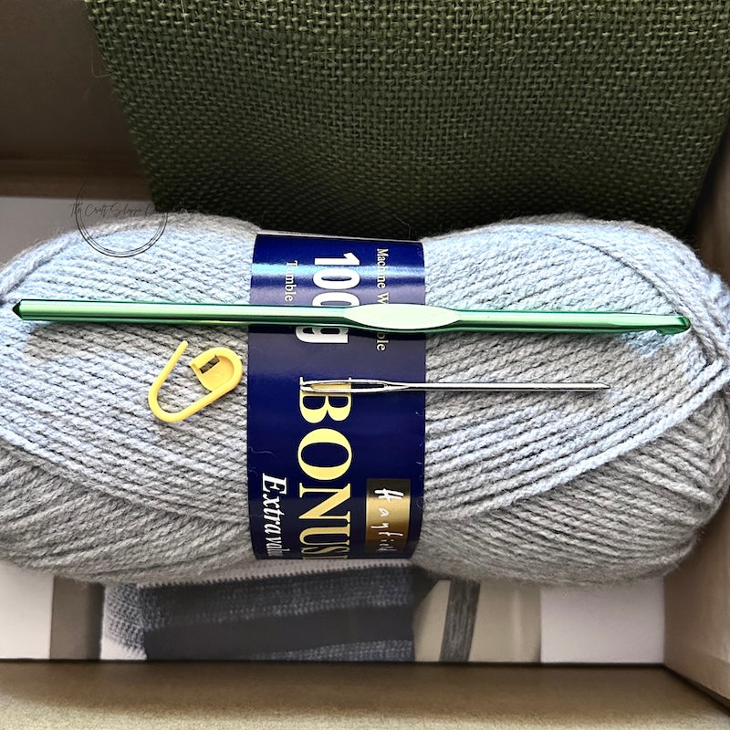 The yarn used in this crochet kit is lying in a box with a crochet hook, stitch marker and a tapestry needle.