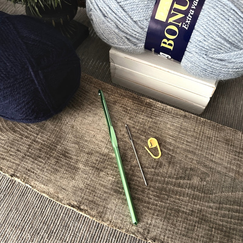 The tools needed to put the couch throw blanket together are lying on a wooden board. There are 2 balls of yarn, a crochet hook, tapestry needle and stitch marker.
