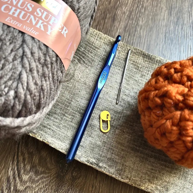 A finished pumpkin, made from this crochet kit, is sitting on a wooden board with yarn and a crochet hook.