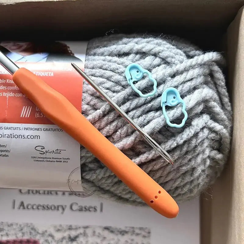 A ball of grey yarn is sitting in a box with a crochet hook, a tapestry needle and 2 stitch markers.