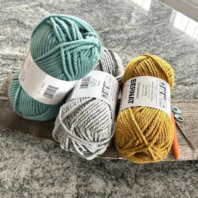 A close up of the teal, grey and yellow yarn, from this crochet kit, on a wooden board with a crochet hook.
