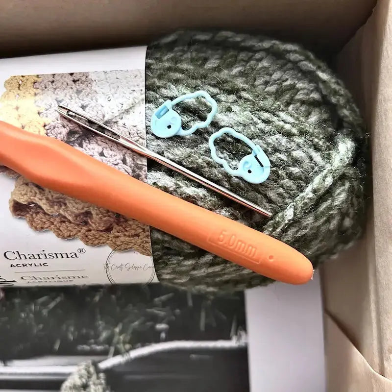 A ball of green yarn is sitting in a box with a crochet hook, a tapestry needle and 2 stitch markers.