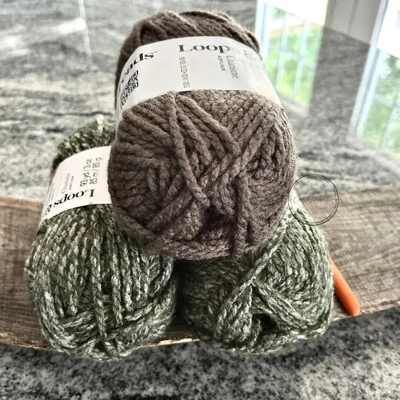 The yarn, that would be used with this crochet pattern, is sitting on a wooden board with a crochet hook.
