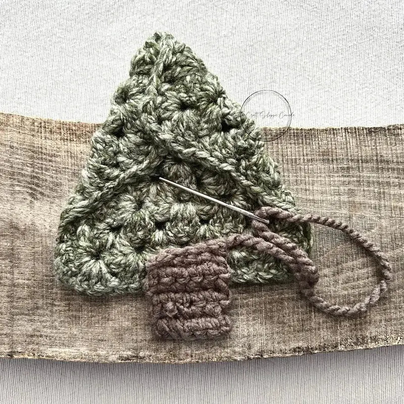 A Christmas tree, made from this crochet pattern, is sitting upside down on a wooden board to show it being stitched with a tapestry needle.