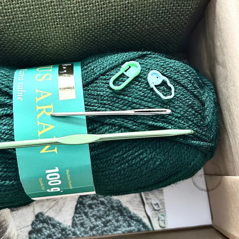 A ball of green yarn is sitting in a box with a crochet hook, a tapestry needle and 2 stitch markers.