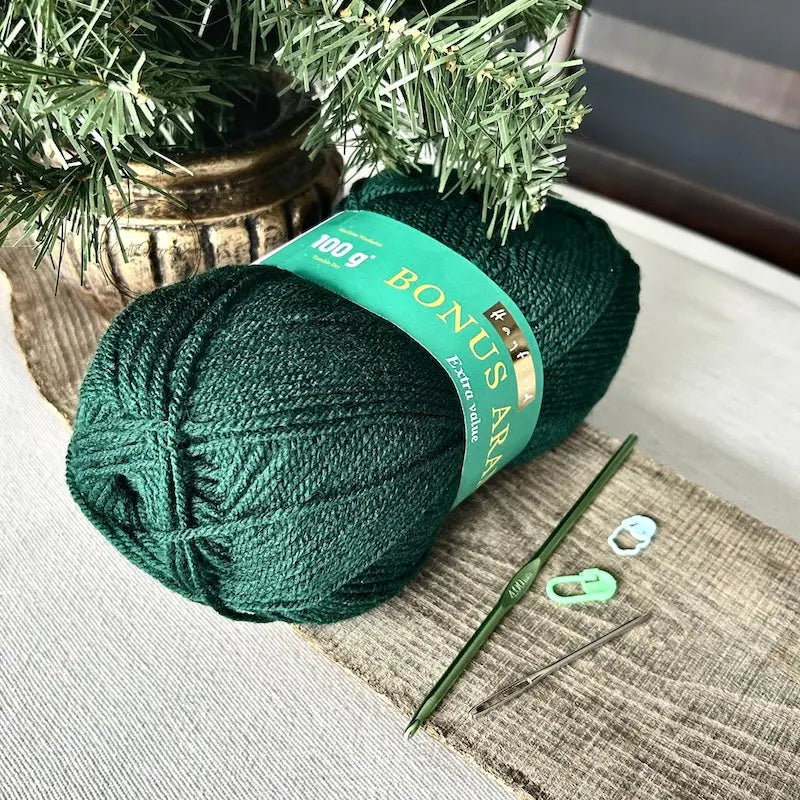 A ball of green yarn, used in this crochet kit is sitting on a wooden board with a crochet hook, tapestry needle and 2 stitch markers.