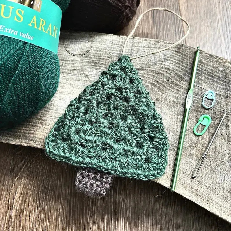 A ball of green and brown yarn, used in this crochet kit is sitting on a wooden board with the finished tree ornament, a crochet hook, tapestry needle and 2 stitch markers.