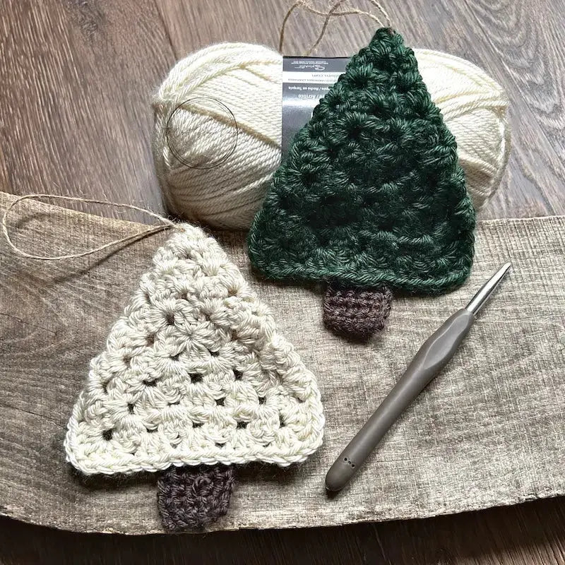 A green and ivory Christmas tree ornament, made from this crochet pattern, are sitting on a wooden board with a crochet hook and yarn.