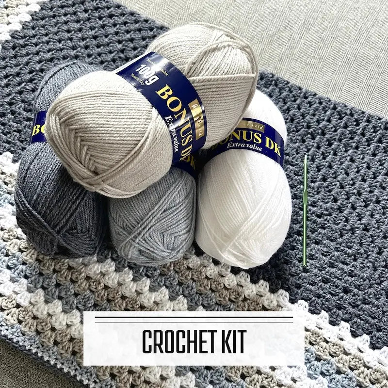 A crochet kit for a granny square blanket. The finished blanket is lying on a couch with 4 balls of yarn and a crochet hook.