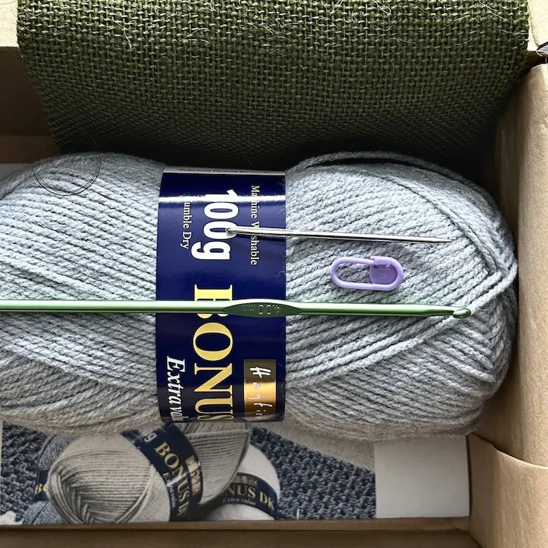 The yarn used in this crochet pattern is lying in a box with a crochet hook, stitch marker and a tapestry needle.