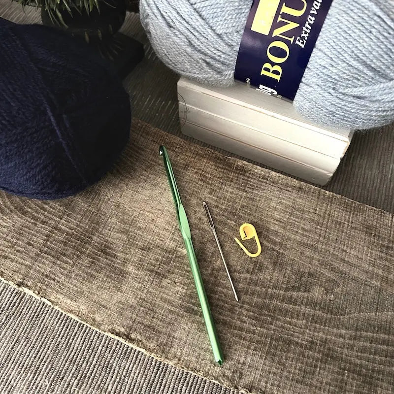 The tools needed to put the couch throw blanket together are lying on a wooden board. There are 2 balls of yarn, a crochet hook, tapestry needle and stitch marker.