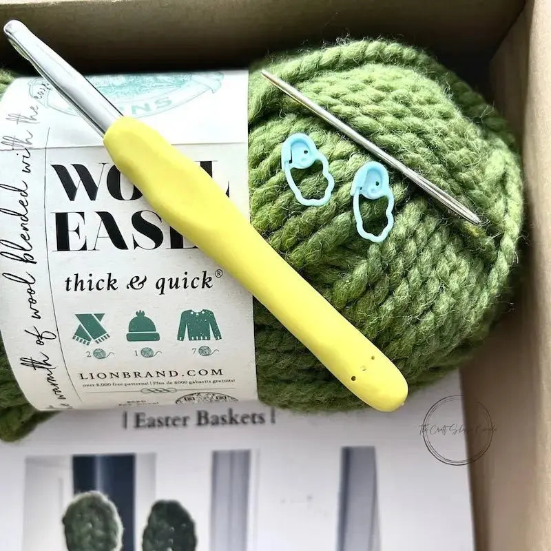 A ball of green yarn is sitting in a box with a crochet hook, a tapestry needle and 2 stitch markers.
