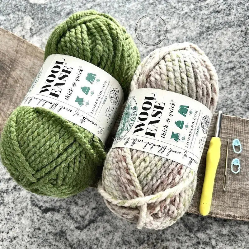 2 balls of yarn lying on a wooden board with a crochet hook, a tapestry needle and 2 stitch markers.