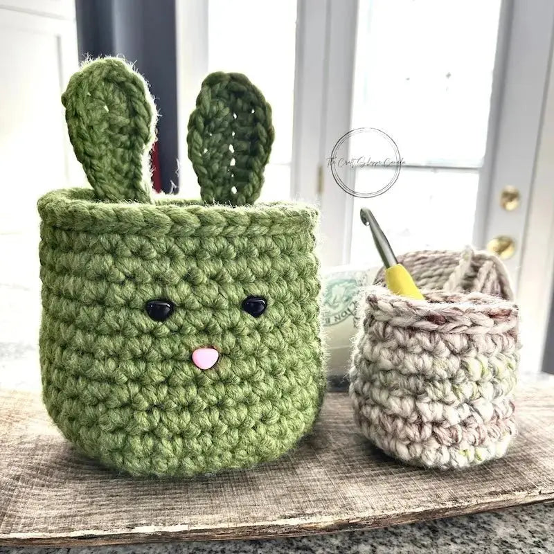 The Easter basket set, from this crochet kit, is sitting on a wooden board with yarn and a crochet hook sitting in the smaller basket.