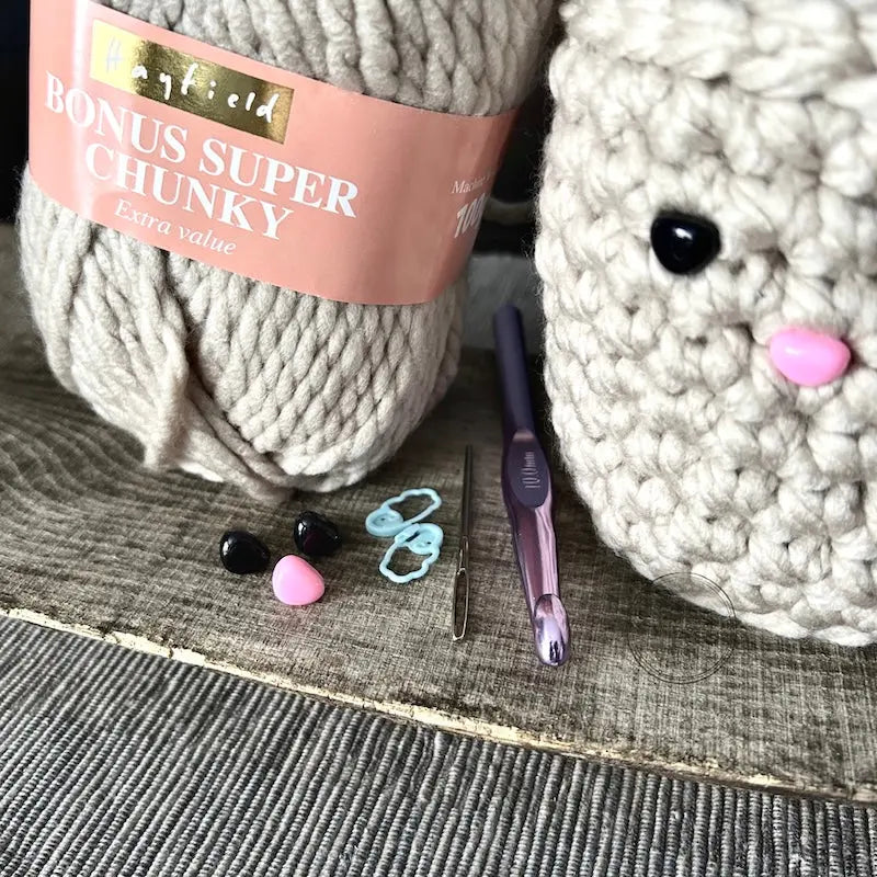 The yarn, crochet hook, tapestry needle, stitch markers and eyes/nose that are included in this crochet kit are sitting on a wooden board.