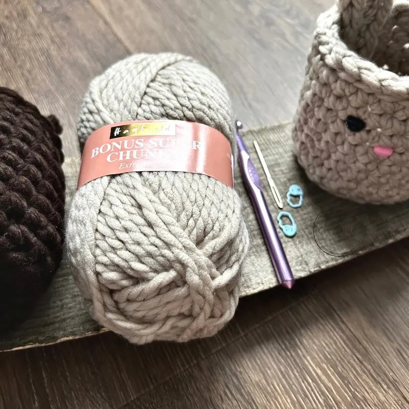 A ball of yarn is lying on a wooden board with a crochet hook, a tapestry needle and 2 stitch markers.