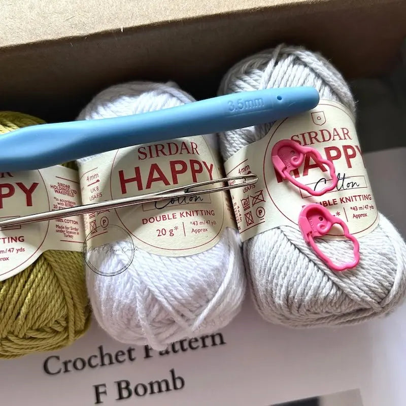 Balls of ivory yarn is sitting in a box with a crochet hook, a tapestry needle and 2 stitch markers.