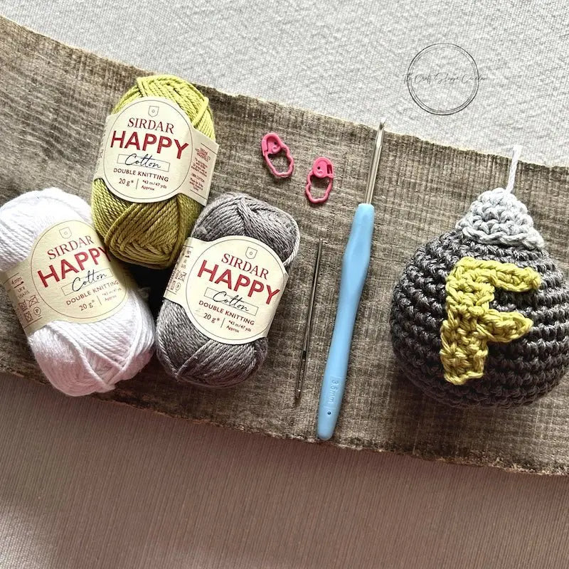 The finished grey F Bomb, made from this crochet kit, lying on a wooden board with yarn, stitch markers and a crochet hook.