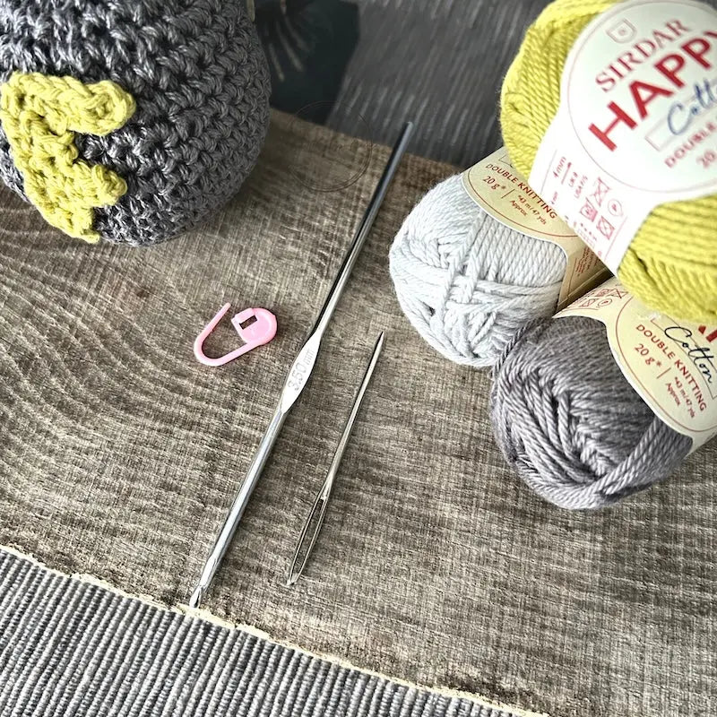 The F-Bomb plush, made from this crochet kit, sitting on a wooden board with a ball of yarn, crochet hook, tapestry and stitch marker.