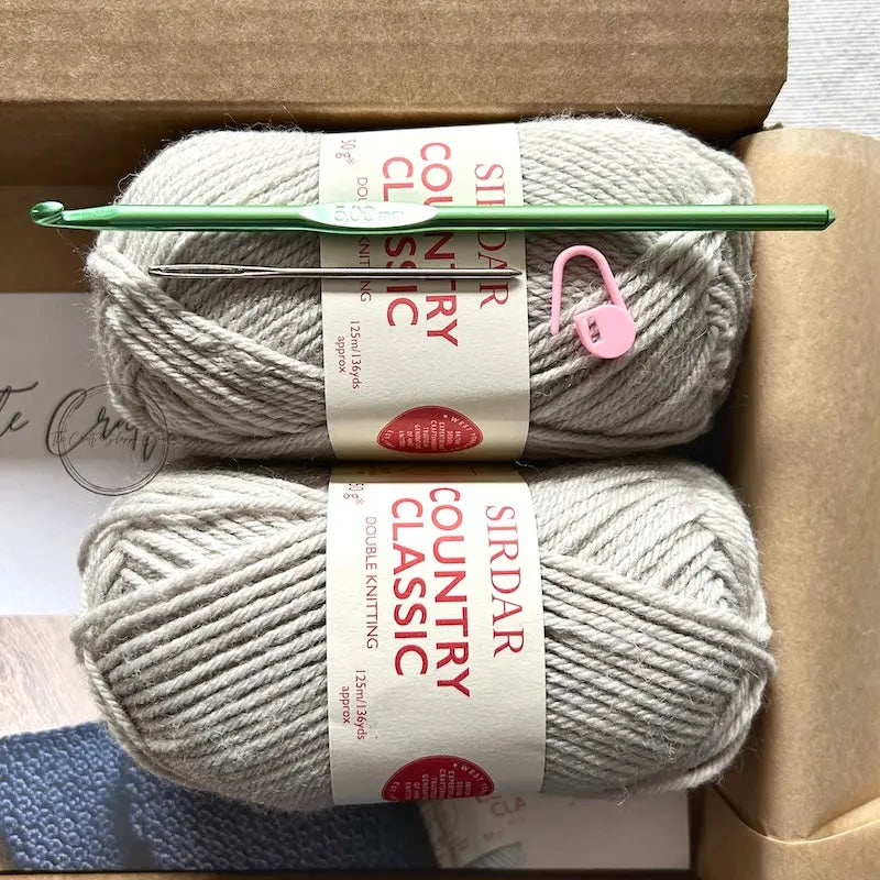 The yarn used in this crochet kit is lying in a box with a crochet hook, stitch marker and a tapestry needle.