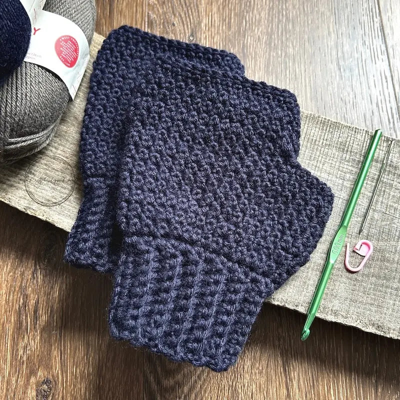 The finished mittens, made from this crochet kit, are lying on a wooden board with yarn and a crochet hook.