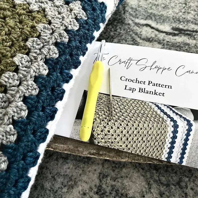 The finished blanket laying on a wooden board, over the crochet pattern with a crochet hook and a tapestry needle.