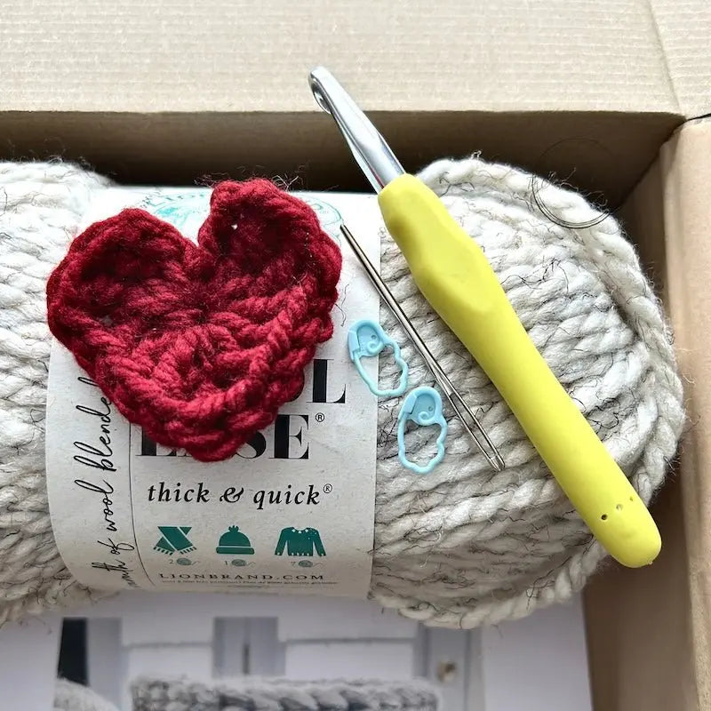 A ball of ivory yarn is sitting in a box with a crochet hook, a tapestry needle and 2 stitch markers.