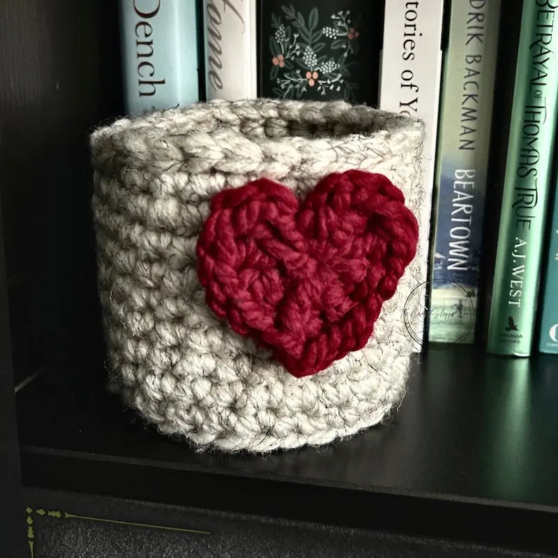 The heart basket, made from this crochet kit, is sitting on a bookshelf.