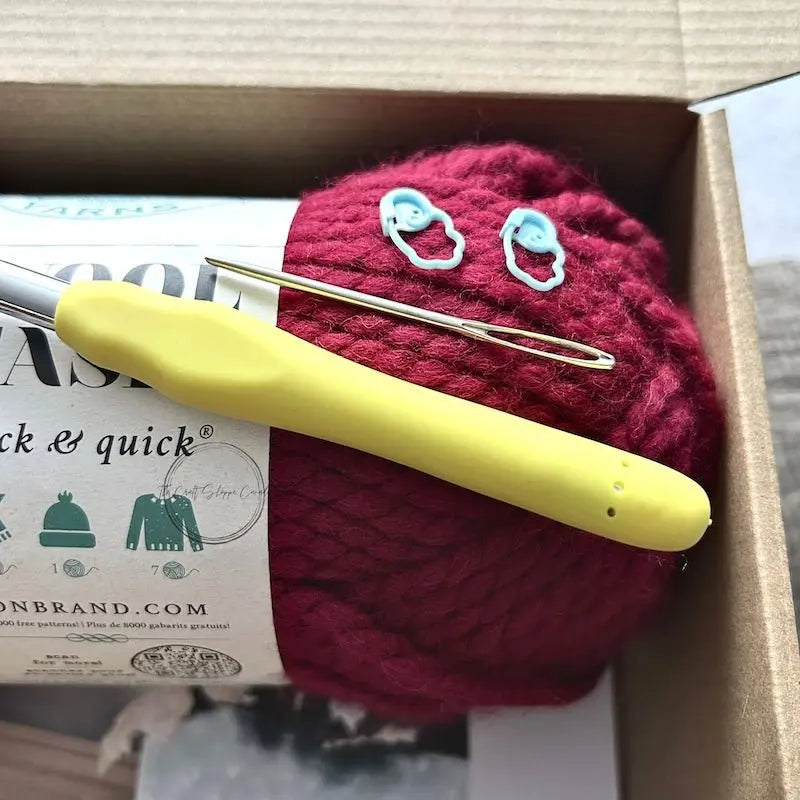 A ball of red yarn is sitting in a box with a crochet hook, a tapestry needle and 2 stitch markers.