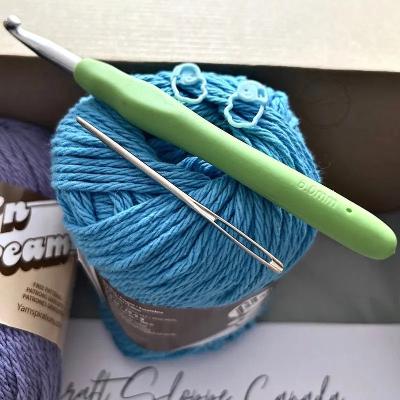 A ball of blue yarn is sitting in a box with a crochet hook, a tapestry needle and 2 stitch markers.