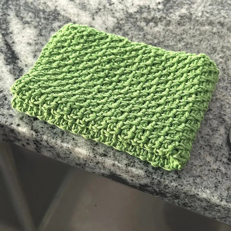 The finished washcloth, made from this crochet kit, is sitting beside a sink.
