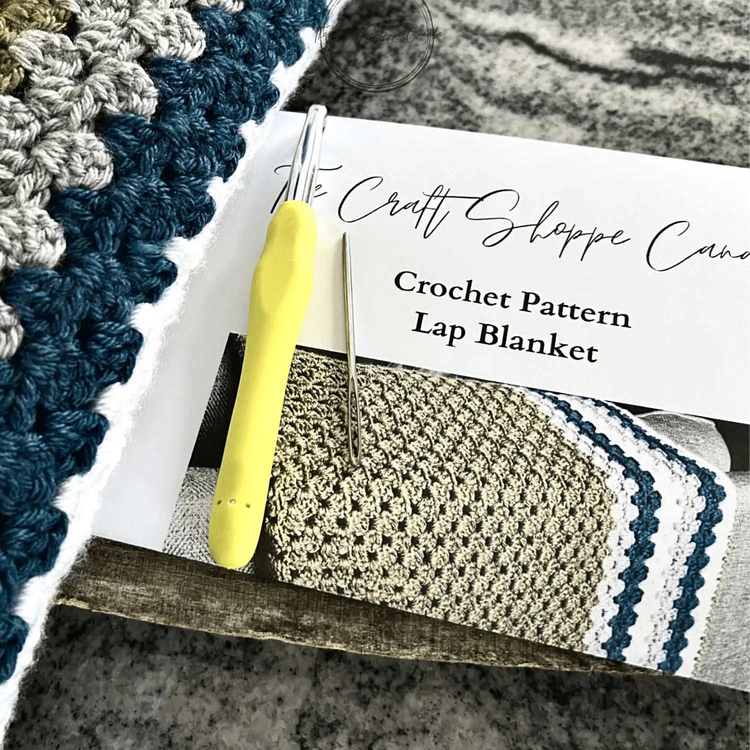 The finished blanket laying on a wooden board, over the crochet pattern with a crochet hook and a tapestry needle.