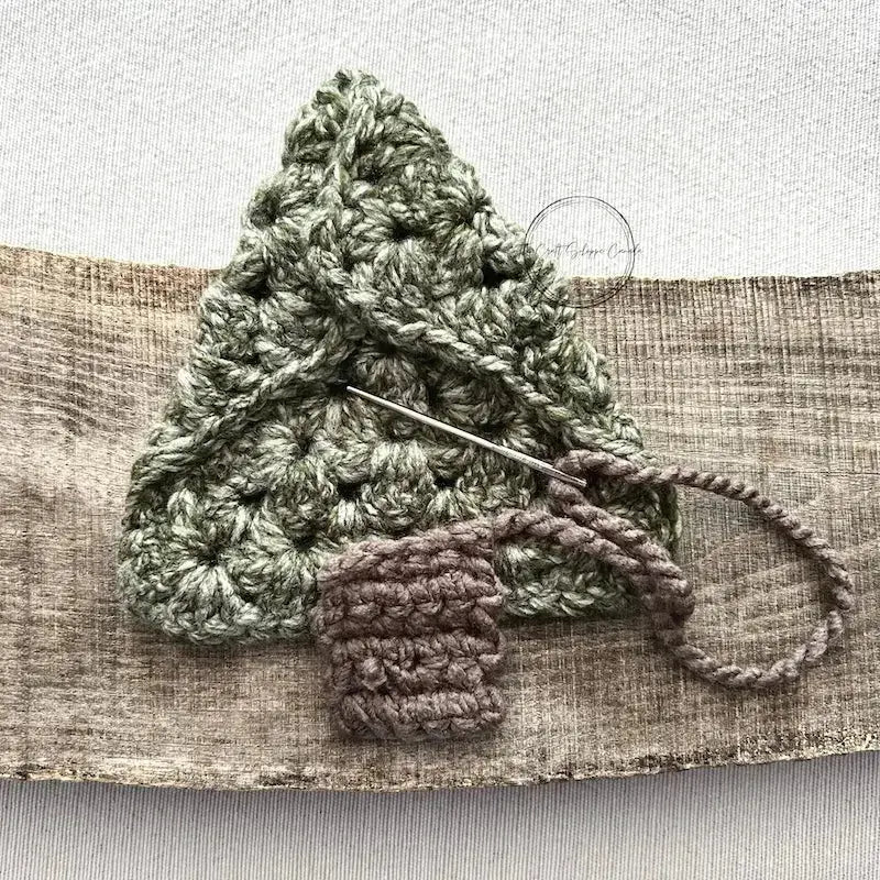 A Christmas tree, made from this crochet pattern, is sitting upside down on a wooden board to show it being stitched with a tapestry needle.
