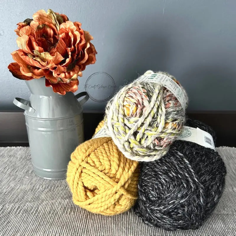 A stack of yarn that you would use in this crochet pattern, are lying on a buffet with a vase of fall flowers beside them.