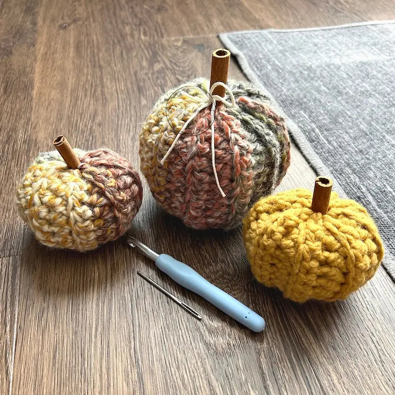3 autumn pumpkins, made from this crochet pattern are sitting on the floor with a crochet hook and tapestry hook in front of them.
