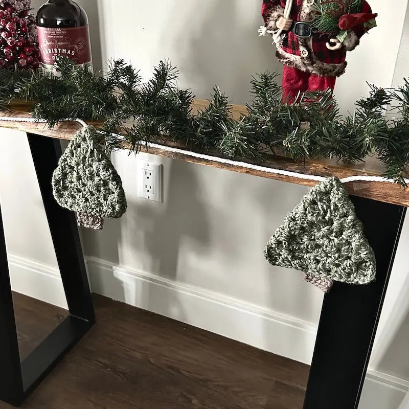 The Christmas garland, made from this crochet pattern, is hanging off of a table with Christmas decorations behind it.