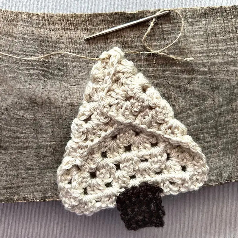 A tree ornament, made from this crochet kit, is sitting on a wooden board. The back of the ornament is facing you with a tapestry needle threading some twine through the top.