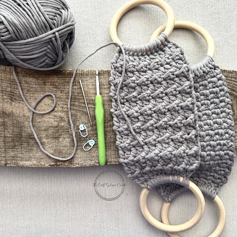 The towel holders, made from this crochet pattern, are lying on a wooden board with yarn, a crochet hook, stitch markers and a tapestry needle.