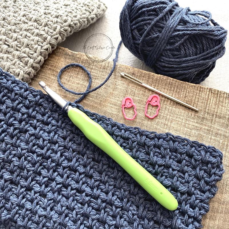The blue and grey dishcloths, made from this crochet pattern, along with yarn, a tapestry needle, stitch markers and a crochet hook.