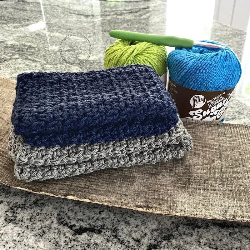 The washcloths, made from this crochet pattern, are folded on a wooden board with balls of yarn and a crochet hook.