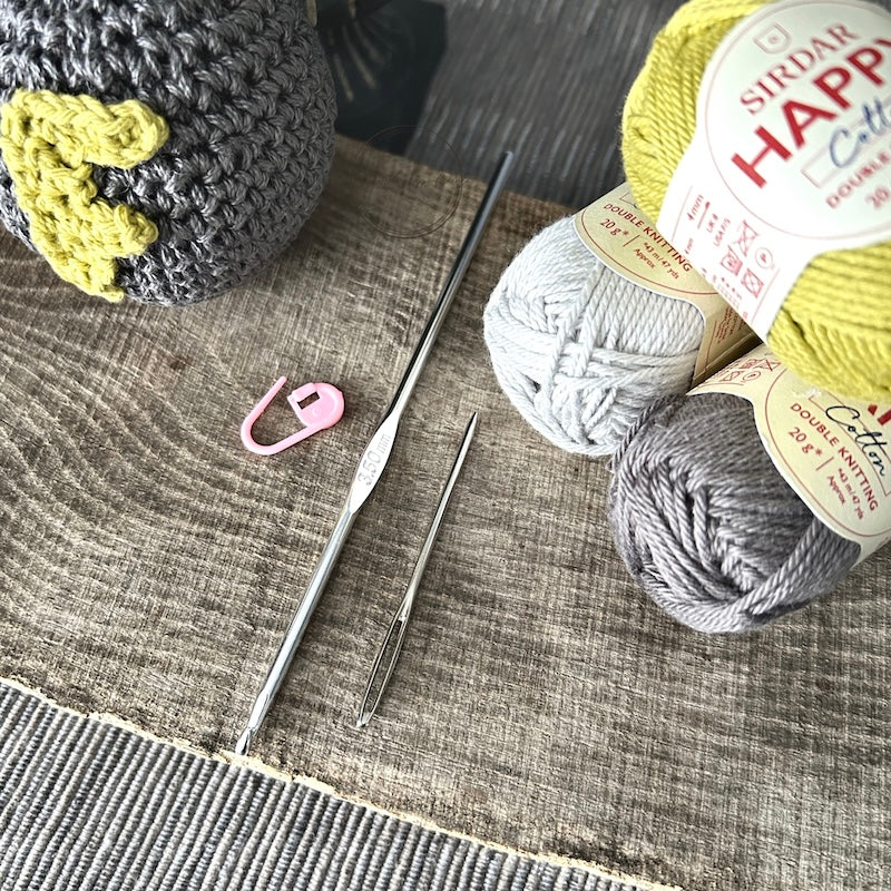 The F-Bomb plush, made from this crochet kit, sitting on a wooden board with a ball of yarn, crochet hook, tapestry and stitch marker.