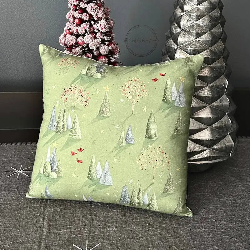 The finished pillow, made from this cross stitch pattern, is sitting backwards on a table against 2 Christmas trees.