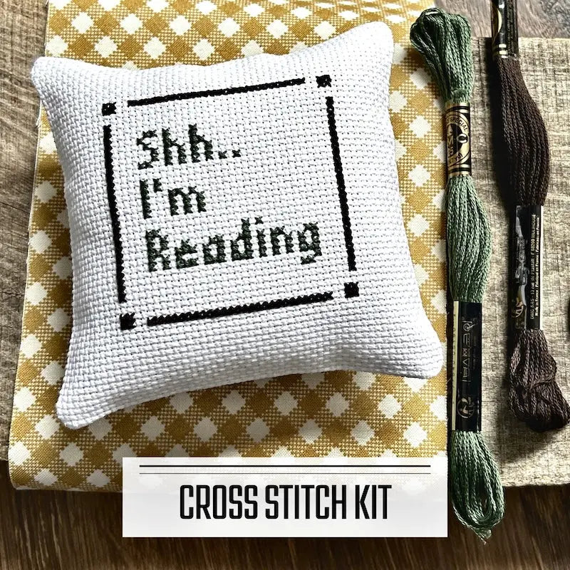 A DIY cross stitch kit for a mini pillow that can be used as a pin cushion. The finished pillow is sitting on a wooden board.