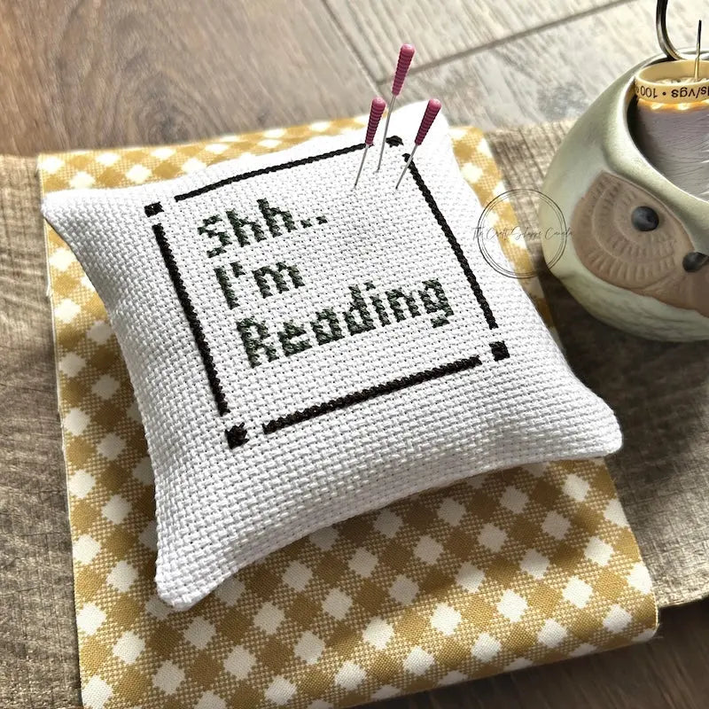 The finished pillow, made from this cross stitch kit, is sitting on a wooden board with pins sticking out.