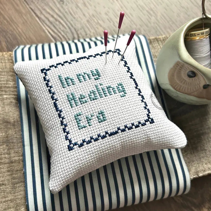 The finished pillow, made from this cross stitch kit, is sitting on a wooden board with pins sticking out.
