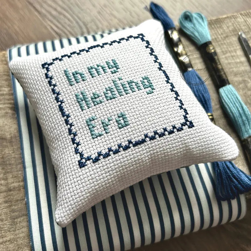 The finished pillow, made from this cross stitch kit, is sitting on a wooden board.