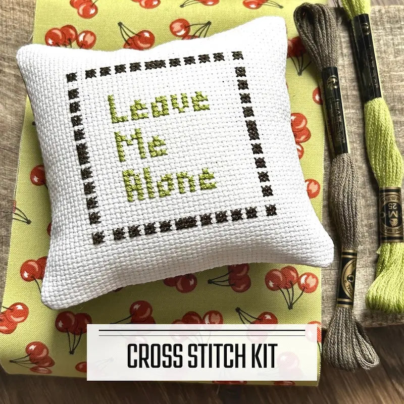 A DIY cross stitch kit for a mini pillow that can be used as a pin cushion. The finished pillow is sitting on a wooden board.
