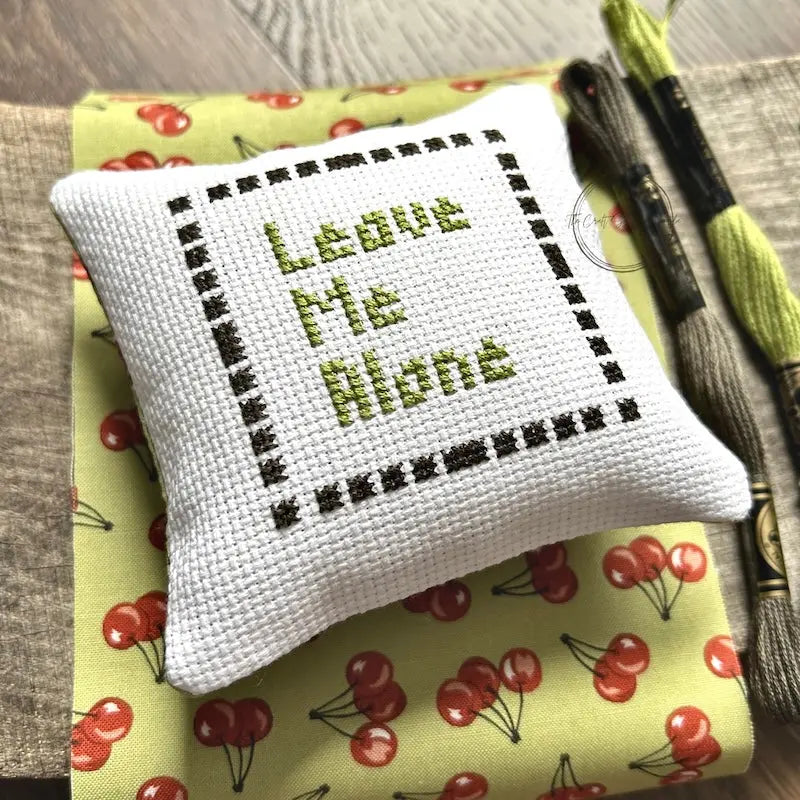 The finished pillow, made from this cross stitch kit, is sitting on a wooden board.